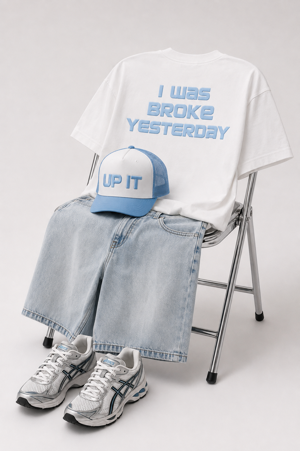 A styled outfit arrangement on a folding chair against a light gray studio background. The set includes a white oversized T-shirt with light blue text reading 'I WAS BROKE YESTERDAY,' light blue denim shorts, a light blue and white trucker cap with 'UP IT' on the front, and white-and-blue athletic sneakers.